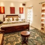 jaisalmer tour packages - Hotel Narpatgarh Palace Jaisalmer Cozy bedroom with a carved wooden bed, white linens, decorative cushions, and elephant-themed curtains.