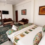 luxury hotels in jaisalmer - Hotel Narpatgarh Palace Jaisalmer Hotel room with a large bed, colorful quilt and pillows, a sofa seating area, wall art of elephants, and a flat-screen TV on the wall.